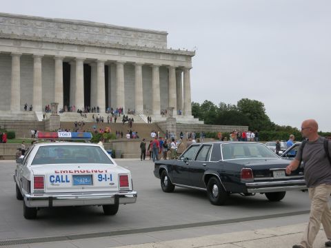 1991 Ford LTD Crown Victoria - white police sedan lettered EMERGENCY CALL 911 and a black sedan parked in front of the Lincoln Memorial with crowds.