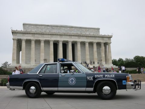 1991 Ford LTD Crown Victoria - navy and silver "90E State Police" Ford sedan sits in side profile in front of the Lincoln Memorial under an overcast sky.