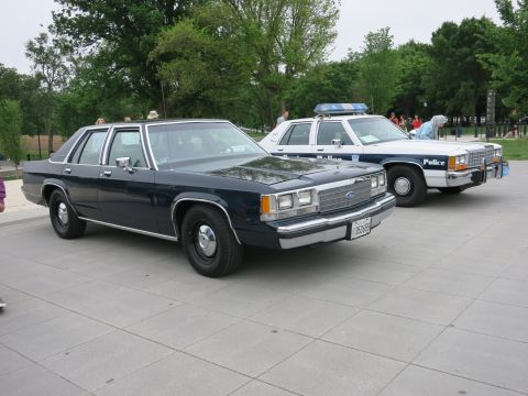 1991 Ford LTD Crown Victoria - dark blue Ford four-door sedan parked on a plaza beside white and blue marked police cruisers under leafy trees.