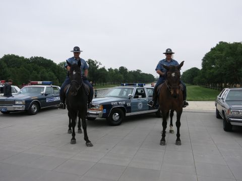 1991 Ford LTD Crown Victoria - Two officers on horseback in dress uniforms pose in front of a navy "90E State Police" Ford cruiser parked among other vehicles on a plaza.