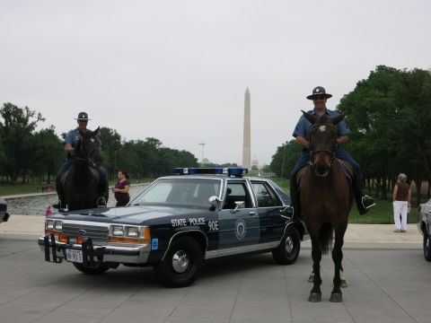 1991 Ford LTD Crown Victoria - Two officers on horseback flank a navy "State Police 90E" Ford cruiser, the Washington Monument rising between them under a gray sky.