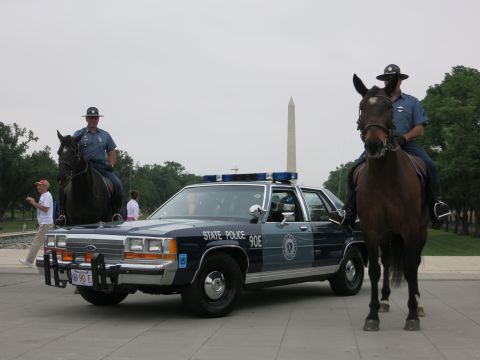 1991 Ford LTD Crown Victoria - Two officers on horseback flank a navy "State Police 90E" Ford cruiser with the Washington Monument behind as bystanders walk past.