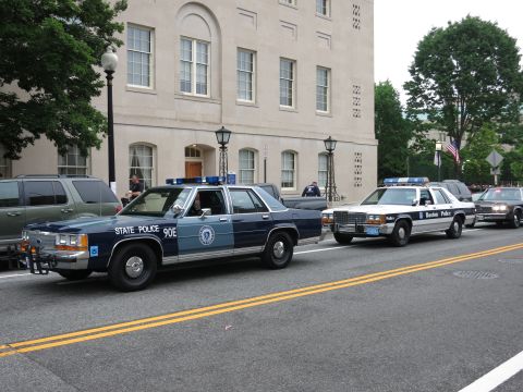 1991 Ford LTD Crown Victoria - navy "90E State Police" Ford cruiser leads a line of marked police cars down a city street beside a stone building flying a U.S. flag.