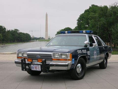 1991 Ford LTD Crown Victoria - navy and silver "State Police 90E" Ford cruiser with push bar viewed from the front three-quarter angle, the Washington Monument behind.