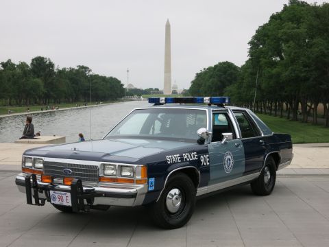 1991 Ford LTD Crown Victoria - Front three-quarter view of a blue-and-silver 'State Police 90E' Ford patrol sedan with roof lightbar parked near the reflecting pool and W…