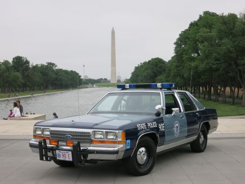 1991 Ford LTD Crown Victoria - Front view of a blue-and-silver 'State Police 90E' Ford patrol sedan with push bar and lightbar, the Washington Monument rising in the hazy…