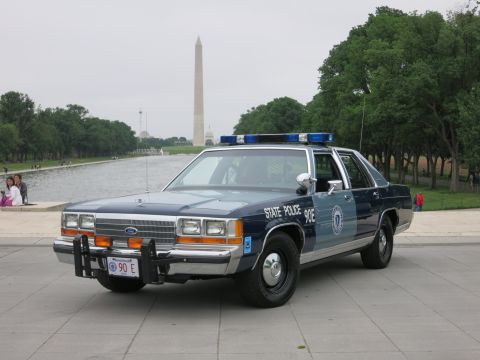 1991 Ford LTD Crown Victoria - blue-and-silver 'State Police 90E' Ford patrol car parked on a stone plaza with the Washington Monument and trees behind under a gray sky.