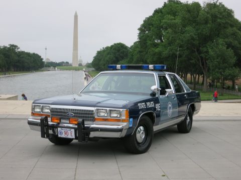 1991 Ford LTD Crown Victoria - Front three-quarter view of a blue-and-silver 'State Police 90E' Ford patrol sedan with push bar and roof lightbar beside the reflecting po…