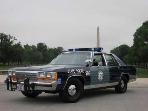 1991 Ford LTD Crown Victoria - blue-and-silver 'State Police 90E' Ford patrol sedan parked on pavement with the Washington Monument centered behind it under an overcast s…