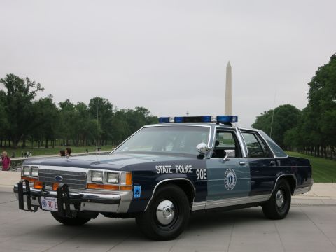 1991 Ford LTD Crown Victoria - blue-and-silver 'State Police 90E' Ford patrol sedan parked on a stone plaza with the Washington Monument and people in the background.