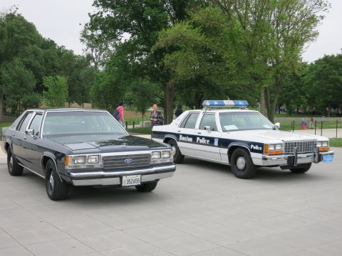 1991 Ford LTD Crown Victoria - black civilian Ford sedan parked next to a white-and-blue 'Boston Police' Ford patrol car with lightbar on a tree-lined stone plaza.