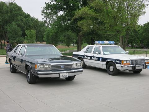 1991 Ford LTD Crown Victoria - black civilian Ford sedan parked beside a white 'Boston Police' Ford patrol car with roof lightbar on a paved plaza among trees.