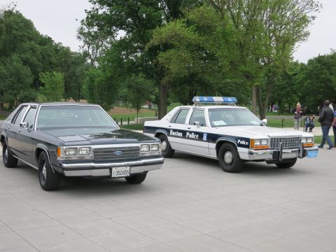 1991 Ford LTD Crown Victoria - black civilian Ford sedan and a white-and-blue 'Boston Police' Ford patrol car parked side by side on a plaza with people walking nearby.