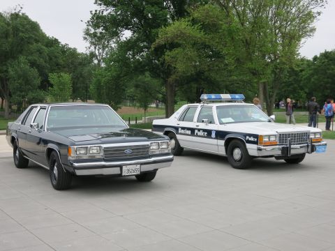 1991 Ford LTD Crown Victoria - black civilian Ford sedan parked next to a white 'Boston Police' Ford patrol car with lightbar on an open paved plaza with onlookers.