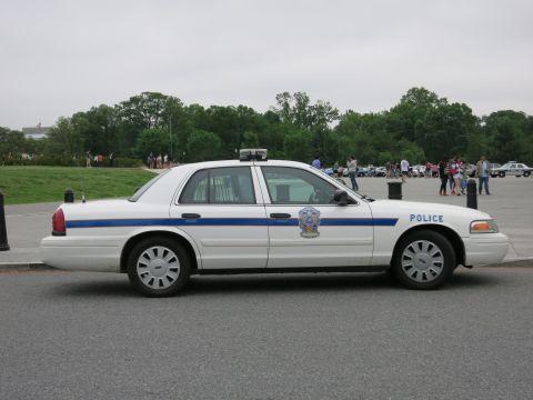 1991 Ford LTD Crown Victoria - Side profile of a white Ford Crown Victoria police sedan marked 'POLICE' with blue stripe, parked on a plaza with trees and crowds behind.