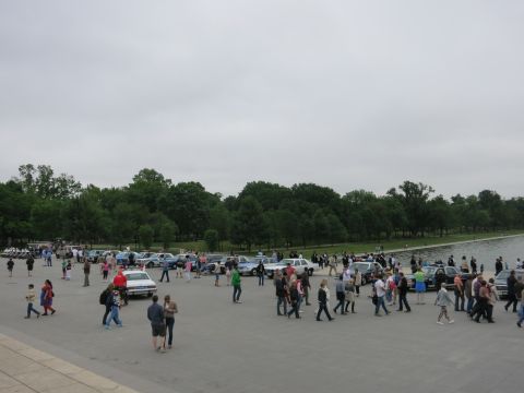 1991 Ford LTD Crown Victoria - large crowd of people walking on a stone plaza near a line of parked vintage cars beside trees and water under an overcast sky.