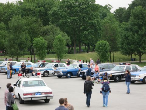 1991 Ford LTD Crown Victoria - parking lot filled with rows of vintage police cars with roof lightbars and spotlights, spectators milling among them by a wooded park.