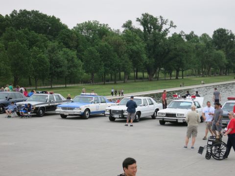 1991 Ford LTD Crown Victoria - Several restored police cruisers are lined up beside a reflecting pool while people gather on the surrounding plaza.