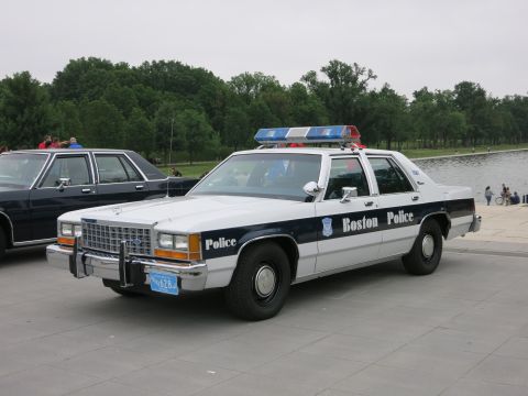 1991 Ford LTD Crown Victoria - restored Boston Police white-over-blue Ford sedan with roof light bar sits parked near a pond on a paved plaza.