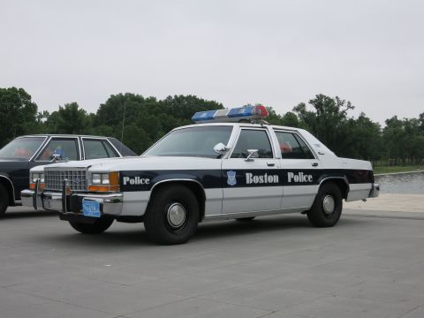 1991 Ford LTD Crown Victoria - white-over-blue Boston Police Ford sedan with roof light bar sits parked on a plaza near a body of water.
