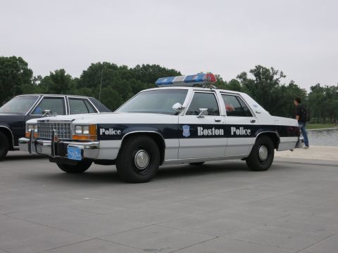 1991 Ford LTD Crown Victoria - restored Boston Police Ford sedan in white and dark blue livery is parked on a stone plaza beside another cruiser.