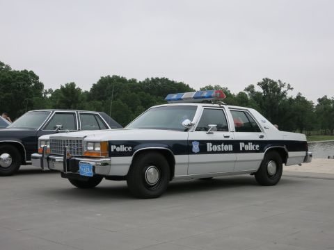 1991 Ford LTD Crown Victoria - Boston Police Ford sedan in white and dark blue colors with a chrome push bar sits parked on a paved plaza near water.