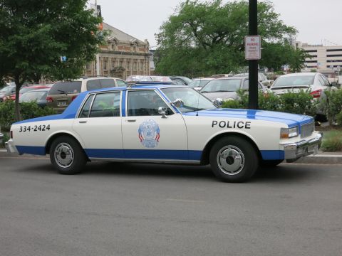 1991 Ford LTD Crown Victoria - white and blue Chevrolet police sedan marked 334-2424 with a department seal is parked along a lot near a stone building.