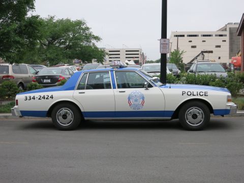 1991 Ford LTD Crown Victoria - white and blue police Chevrolet sedan with the number 334-2424 and a department seal is parked along a street.