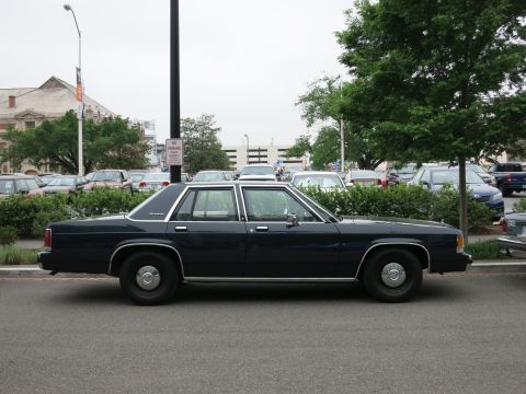 Side view of a dark blue full-size sedan parked in front of shrubs and a parking lot