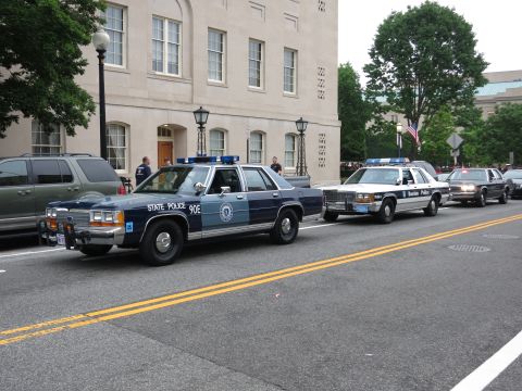 Blue Massachusetts State Police sedan leading other cruisers on a street