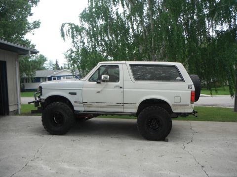 1990 Ford Bronco - White 1990 Ford Bronco lifted with large off-road tires sits on a concrete driveway.