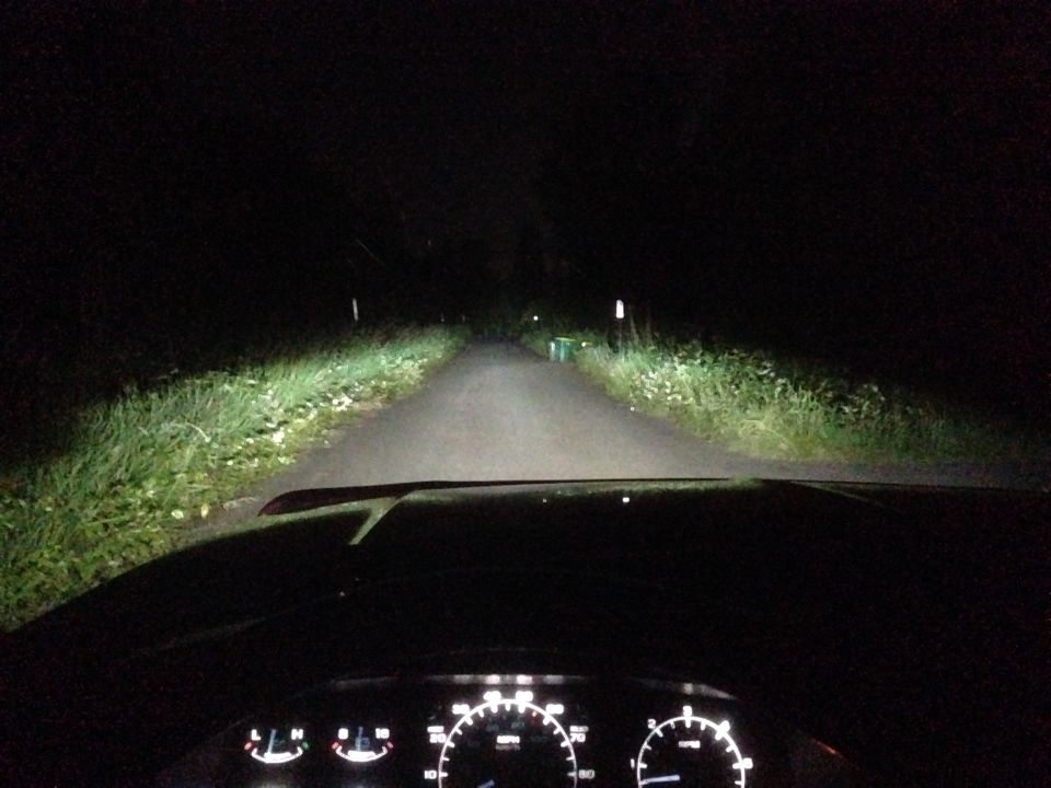 Dashboard view at night, showing illuminated gauges and bright headlights illuminating a rural road