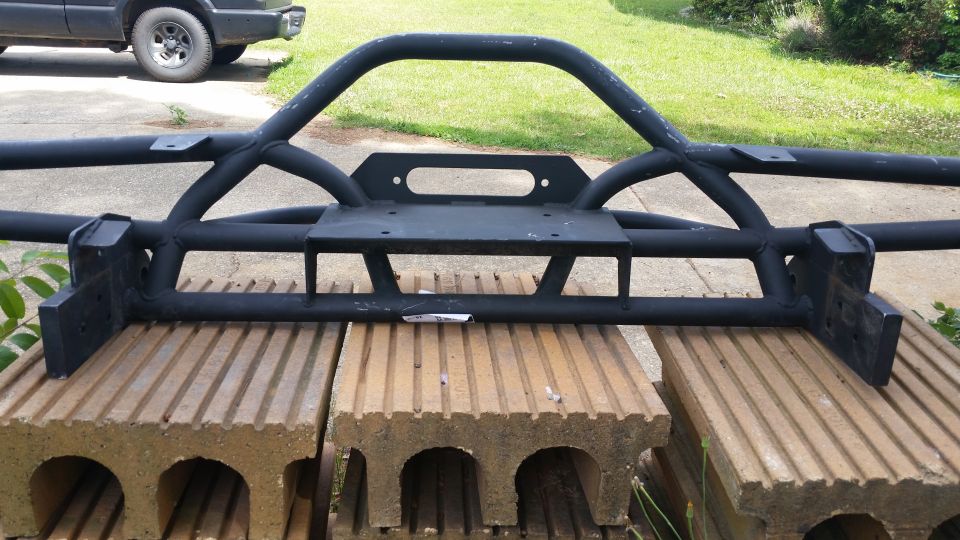 black steel off-road bumper with winch mount, sitting on concrete blocks. Part of a dark SUV visible in background