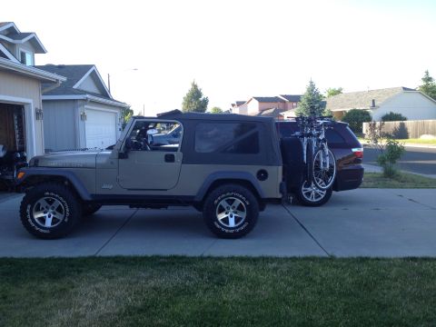 2005 Jeep Wrangler - A tan Jeep Wrangler is parked in a driveway, with a dark SUV and bicycles visible behind it.
