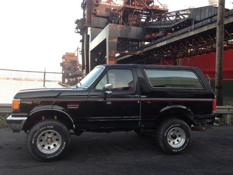 1988 Ford Bronco - A black Ford Bronco is parked outdoors with industrial equipment in the background.