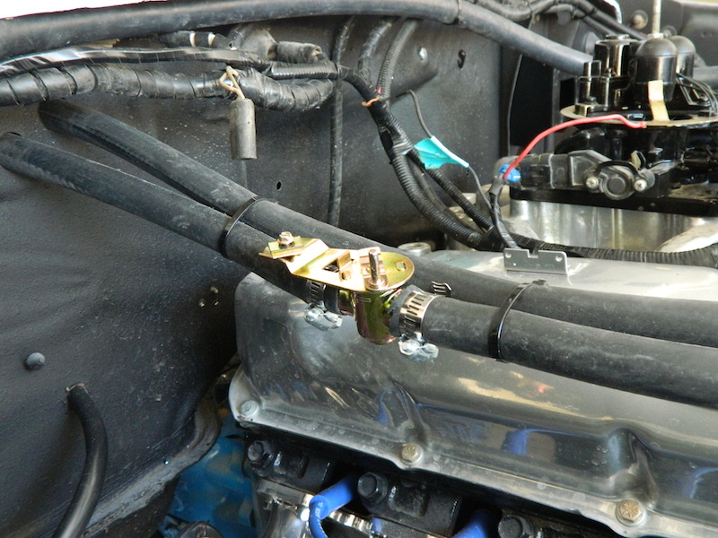 Engine bay showing two black hoses connected by a metal bracket near a carburetor