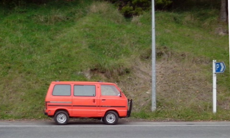 Orange van parked on the side of the road next to a grassy hill and a parking sign