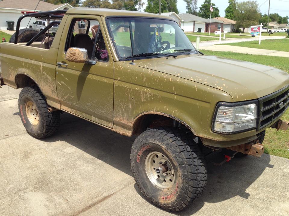 Lifted olive-green Ford Bronco with top off, roll bar, mud splatter, and large General Grabber tires in a driveway
