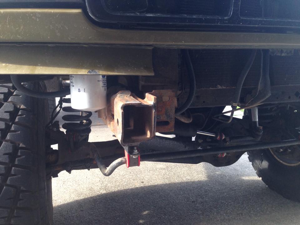 Underside front of lifted Bronco showing rusty receiver hitch tow point, white filter, coil springs and steering linkage