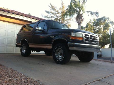 1994 Ford Bronco - A dark blue Ford Bronco is parked in front of a garage with a palm tree in the background.
