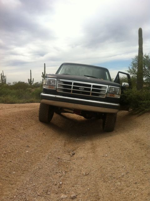 1994 Ford Bronco - A dark-colored Ford F-Series truck is parked on a dusty trail, with saguaro cacti in the background under a cloudy sky.