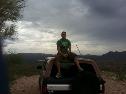 1994 Ford Bronco - Man sits atop a 1994 Ford Bronco, enjoying a vast mountain vista under a cloudy sky.