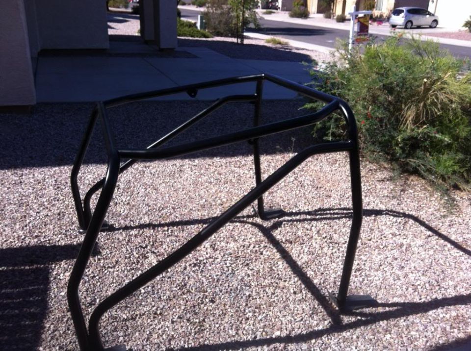 Black tubular roll cage with foot plates sitting on gravel outside a house