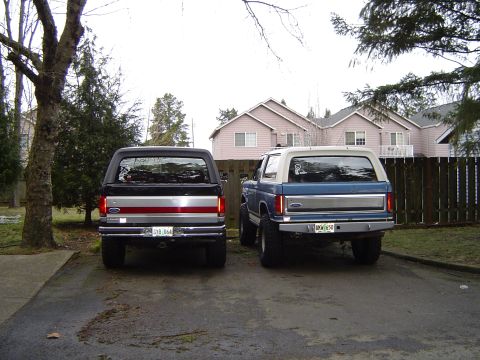 1989 Ford Bronco - Two Ford Broncos, one blue and one black, are parked in a driveway, viewed from the rear.