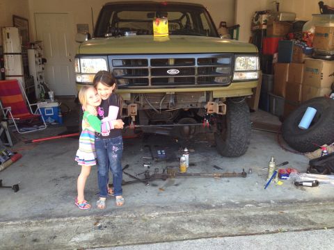 1994 Ford Bronco - Two young girls embrace in front of a green 1994 Ford Bronco undergoing repairs in a garage.