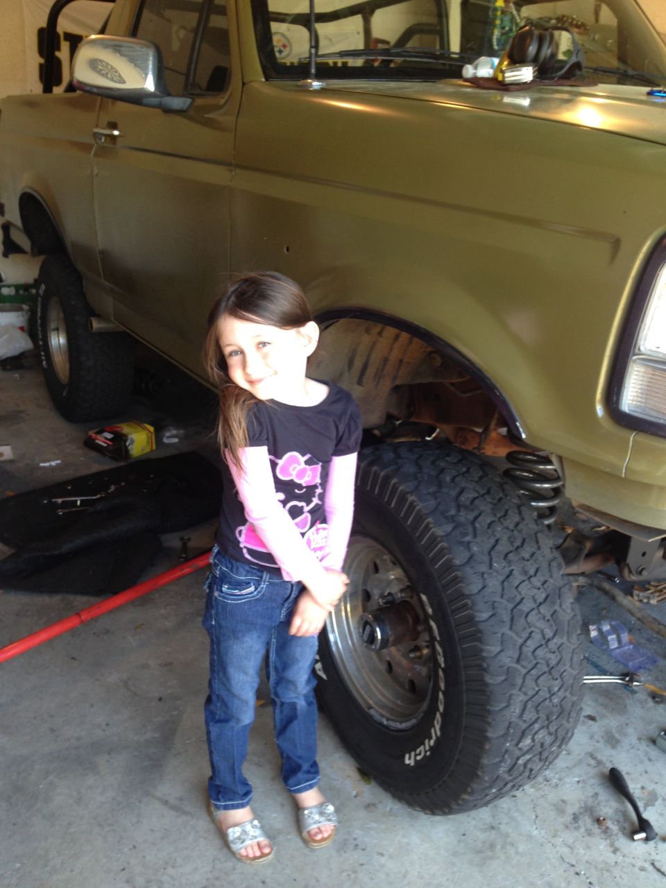 Girl in front of a lifted, olive-green truck that appears to be from the early 90s. The truck is in a garage, possibly undergoing maintenance