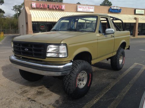 1994 Ford Bronco - Olive green 1994 Ford Bronco with aftermarket roll cage and large tires is parked in front of a strip mall.