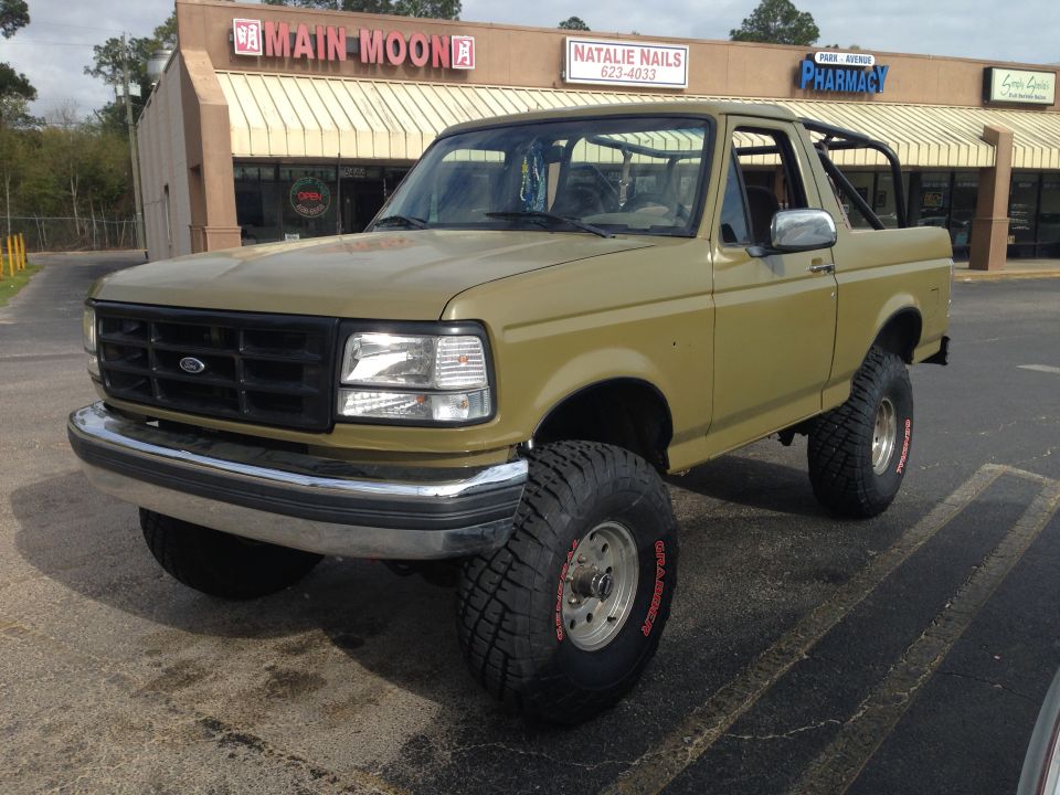 Olive green 1994 Ford Bronco with aftermarket roll cage and large tires parked in front of a strip mall