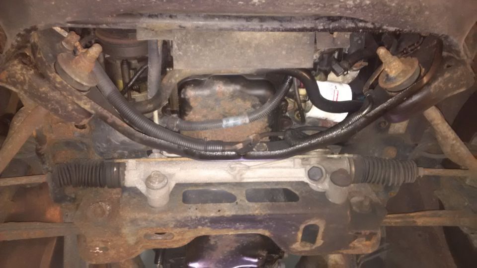 Underside view of a vehicle's steering rack and engine oil pan. Lots of rust and grime visible