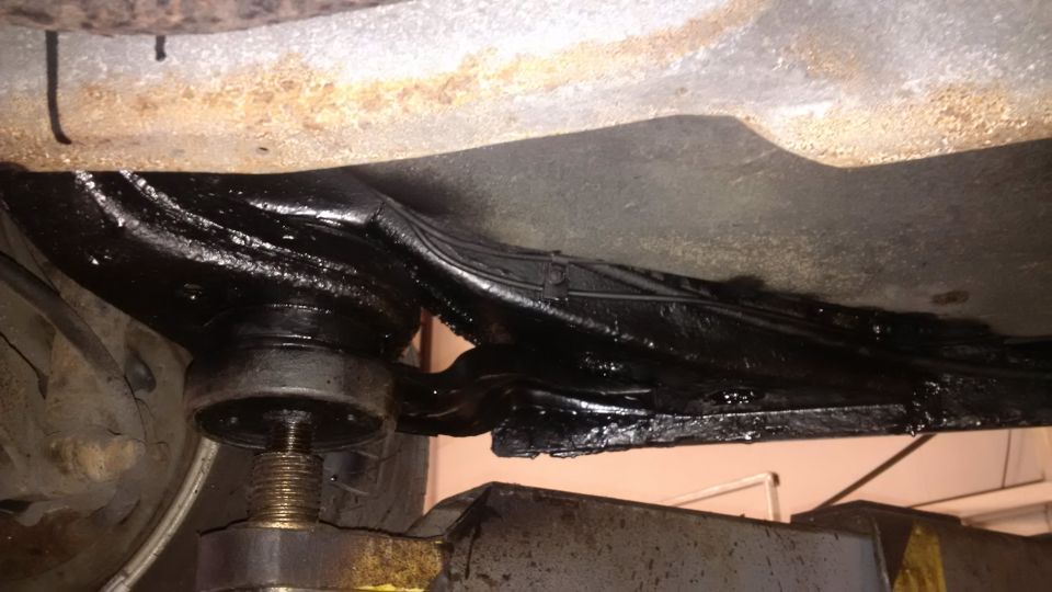 Undercarriage of a vehicle with black undercoating applied to the frame and suspension components. A jack is visible supporting the frame